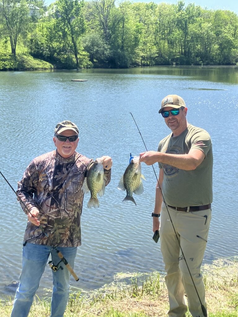 Two guests standing at the edge of the stocked pond at Selah Hills Ranch holding up the crappie they caught, rods still in hand.